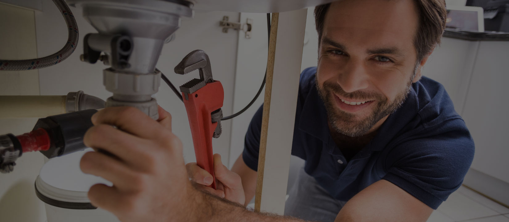 Smiling Brisbane plumber using a pipe wrench to tighten under-sink plumbing connections in a kitchen cabinet