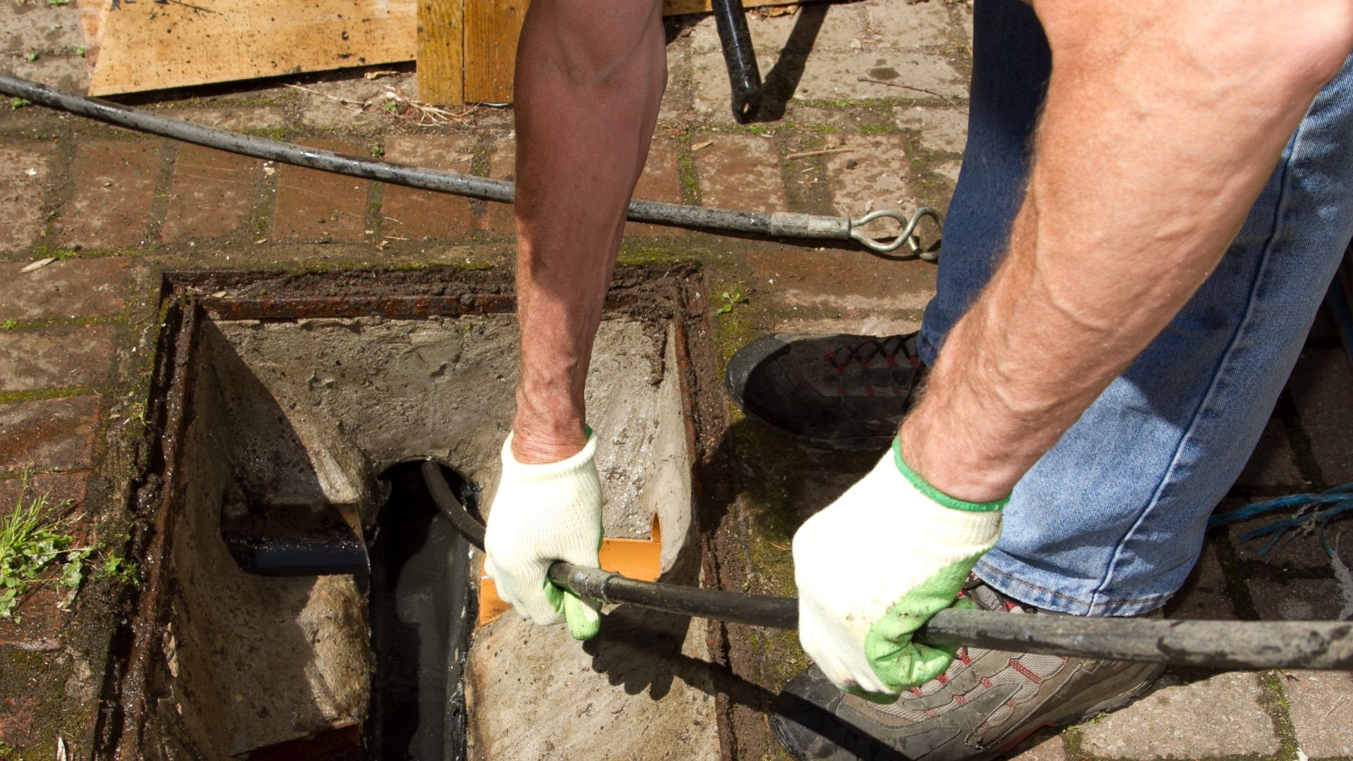 The image shows a plumber unclogging a drain with a plumbing tool while wearing gloves, standing over a sewer drain.