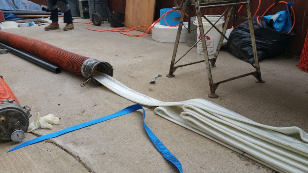 A person is unrolling a large blue hose from a coiled container on the ground. The scene shows a construction or plumbing area with tools and materials scattered around. In the background, there is a metal frame, bags, and other equipment