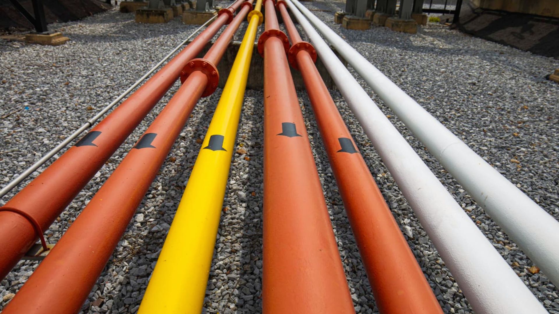 Rows of colorful industrial pipes (red, yellow, and white) placed on gravel ground with black directional arrows marked along their length.