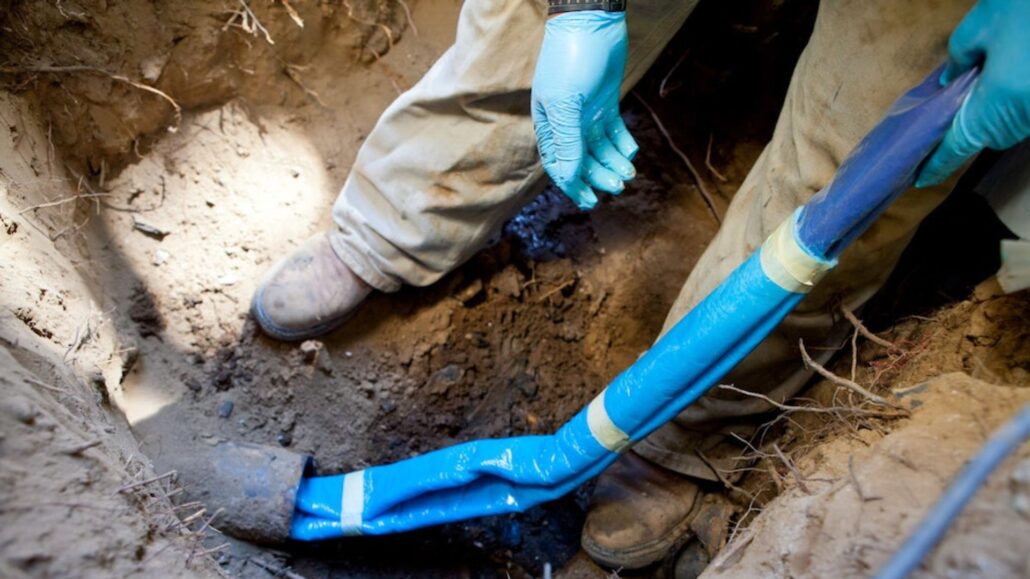 A plumber in protective gloves works in a dug trench, holding a piece of blue pipe wrapped in tape, possibly performing a repair or installation on an underground water line.