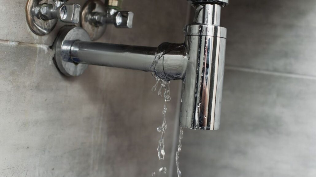 A close-up image of a leaking chrome pipe joint, with water dripping from it, set against a grey tiled wall.