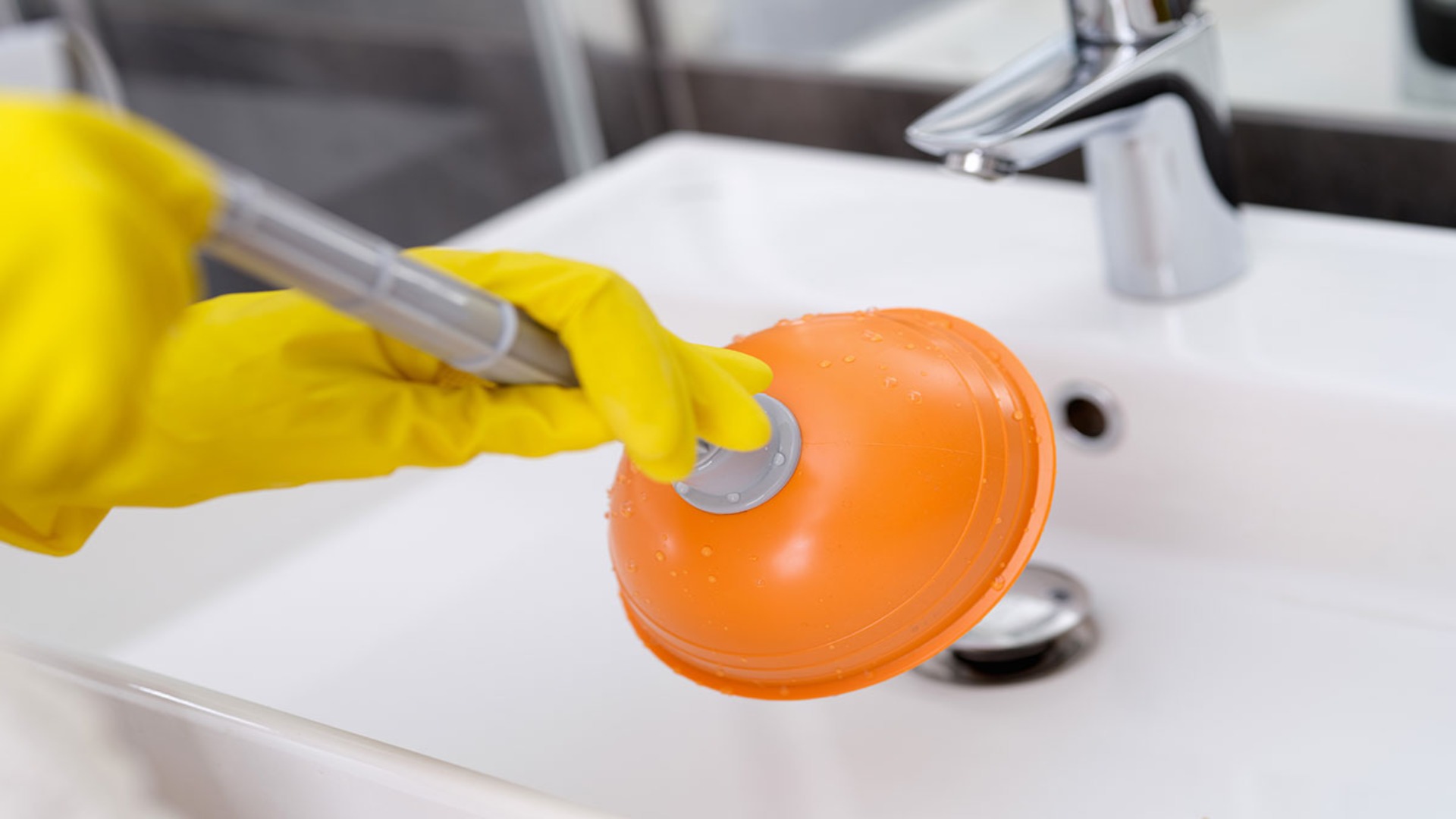 A person wearing yellow rubber gloves is using a plunger on a sink to unclog it. The sink is clean, and the plunger is orange.