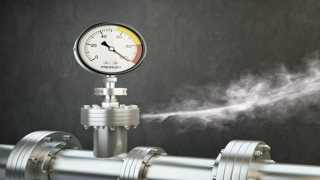 A close-up of a pressure gauge on a pipe, showing pressure levels with steam escaping from the pipe.