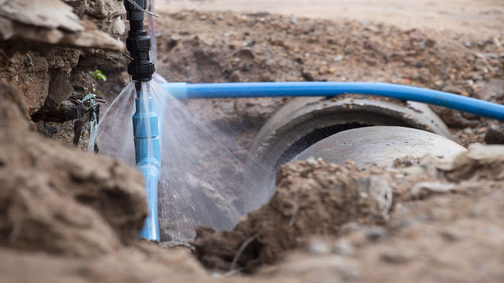 Underground water pipe leaking from a cracked blue service line, with water spraying at a construction excavation site, illustrating emergency pipe repair and leak detection services offered by Brisbane plumbers.