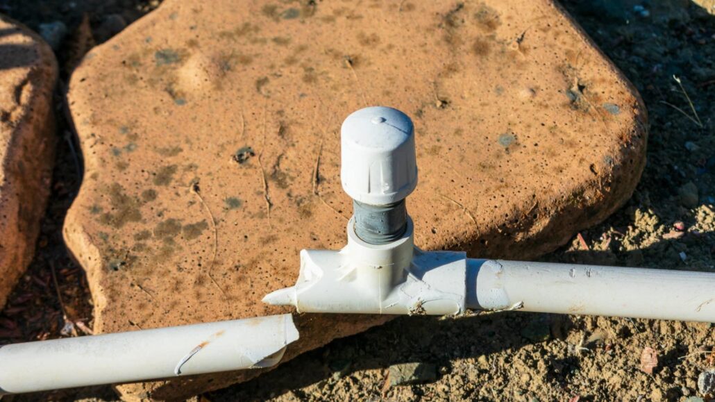 A close-up image of a cracked white PVC pipe with a valve cap installed, lying on a dirt surface near a stone.