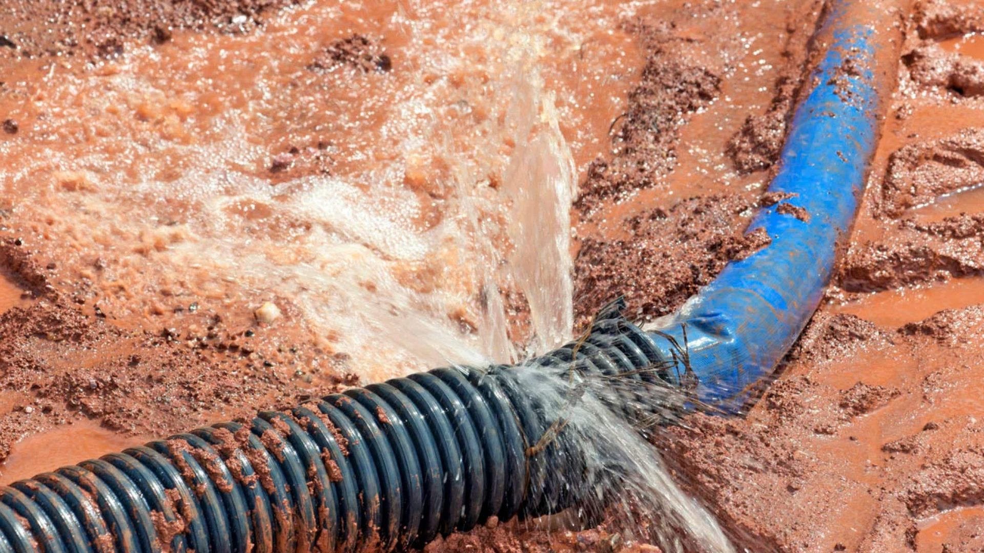 A damaged blue hose spraying water with mud around it, showing signs of leakage at the point of breakage.