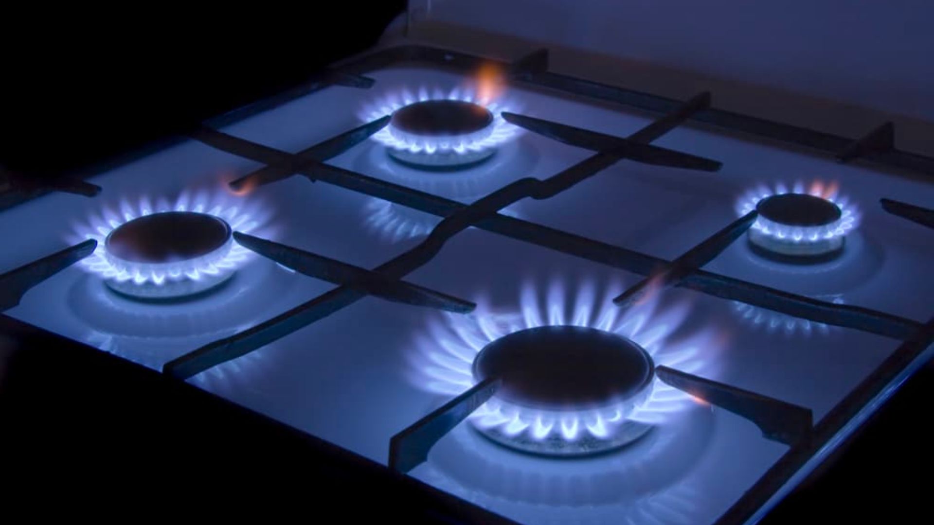 A close-up of a gas stove with four burners, all emitting blue flames in a dark setting, showing active cooking zones.