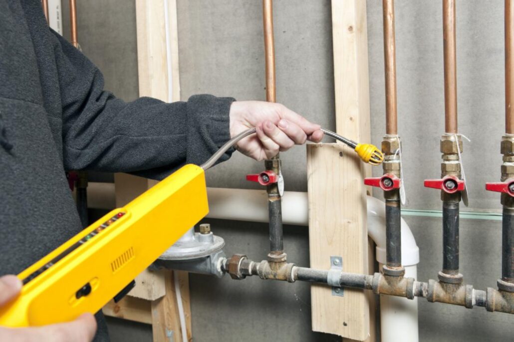 A person is using a yellow gas leak detector to check copper pipes with red valve handles in a plumbing setup.