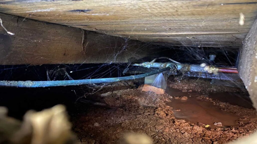 The image shows a water pipe leaking beneath a wooden floor, with water spraying from a break in the pipe. The surrounding area is muddy, and spider webs are visible near the pipe.
