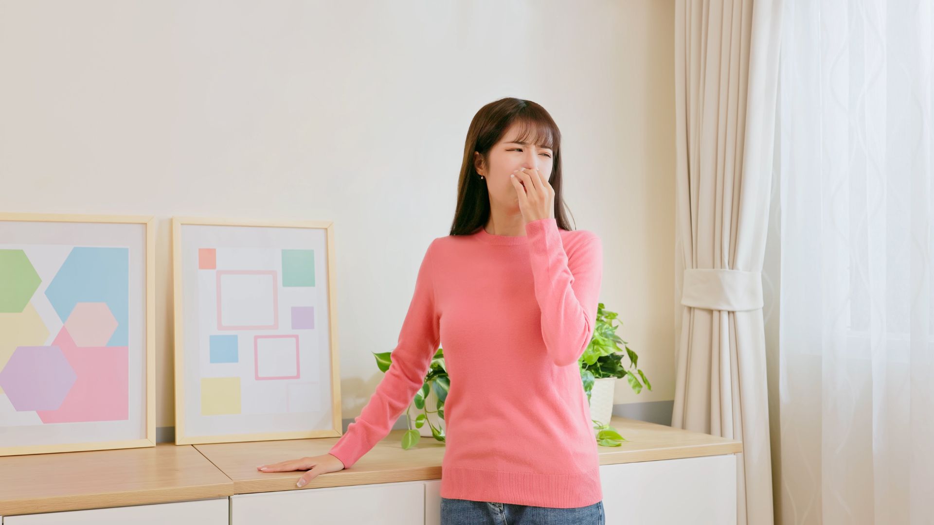 Woman in a pink sweater standing in front of colorful framed artwork, holding her nose and reacting to an unpleasant smell, in a bright room with plants and curtains.