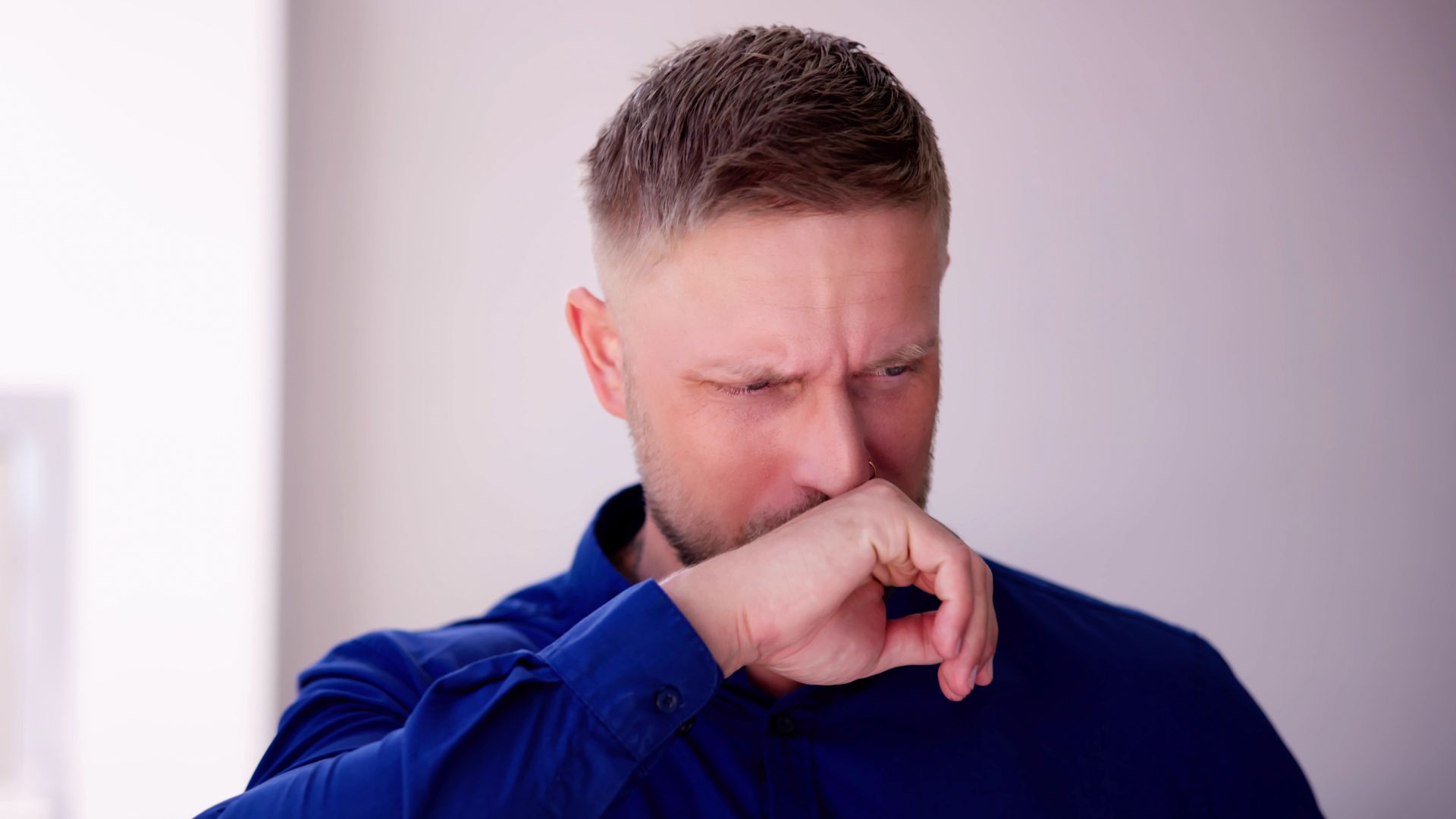 Man in a blue shirt sniffing and holding his nose, indicating a reaction to a bad smell in a minimalistic indoor setting.
