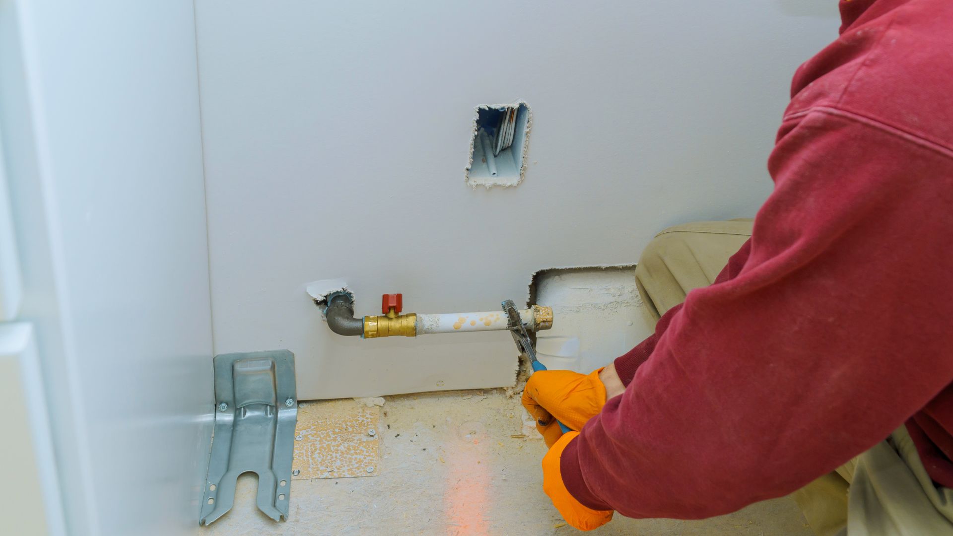 Plumber wearing gloves using a wrench to tighten or fix a pipe, with a wall-mounted plumbing setup and exposed pipes in a laundry room.