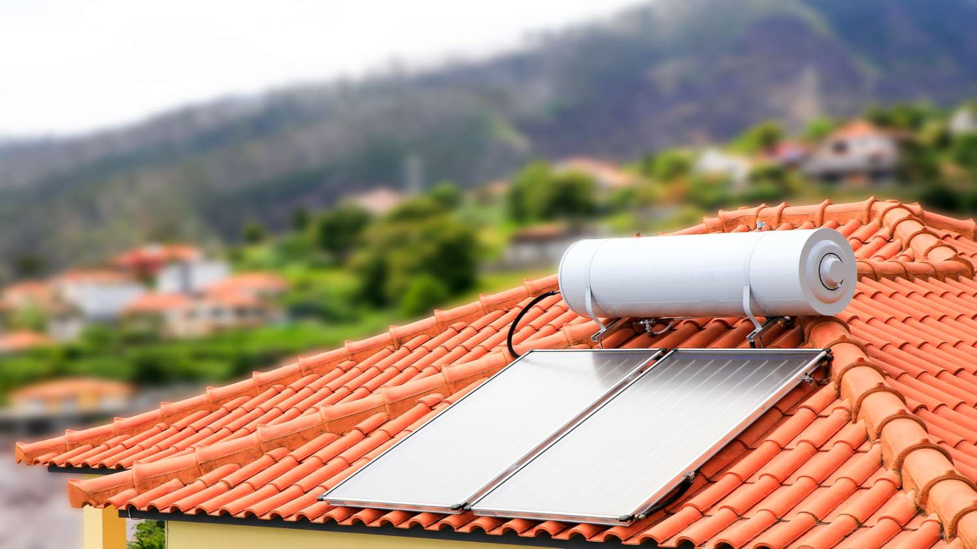 A close-up image of a solar water heater system installed on a red-tiled roof. The solar panels are positioned below a cylindrical water tank, with a scenic background of houses and greenery.