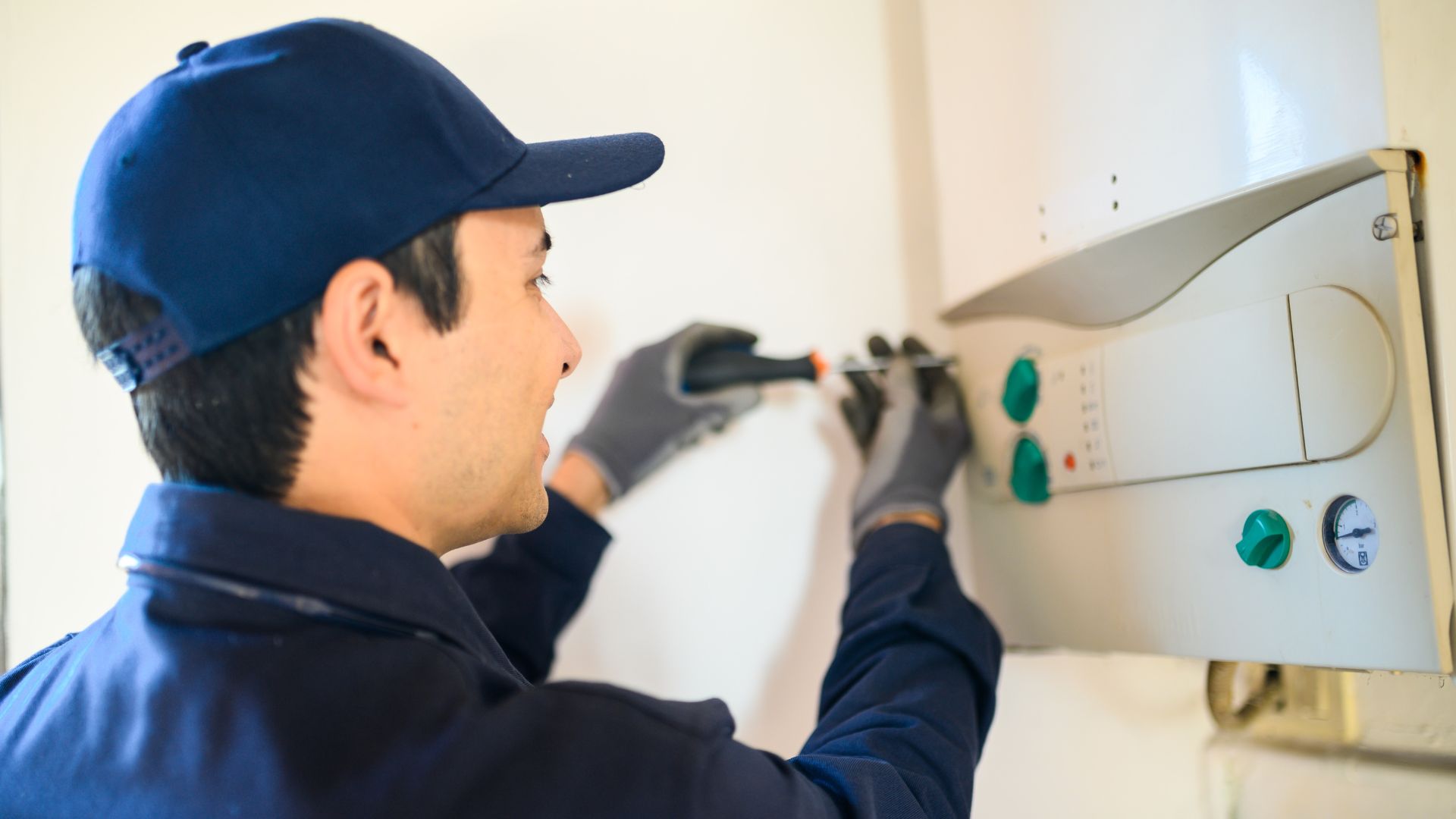 Plumber in a blue uniform using a screwdriver to adjust the controls on a water heater mounted on the wall.