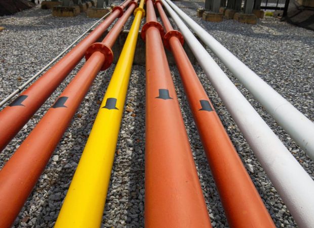 Rows of colorful industrial pipes (red, yellow, and white) placed on gravel ground with black directional arrows marked along their length.