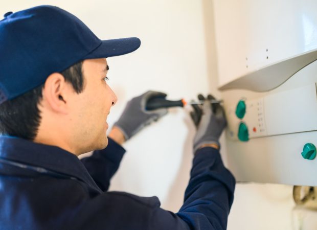 Plumber in a blue uniform using a screwdriver to adjust the controls on a water heater mounted on the wall.