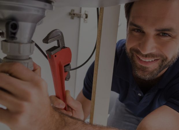 Smiling Brisbane plumber using a pipe wrench to tighten under-sink plumbing connections in a kitchen cabinet