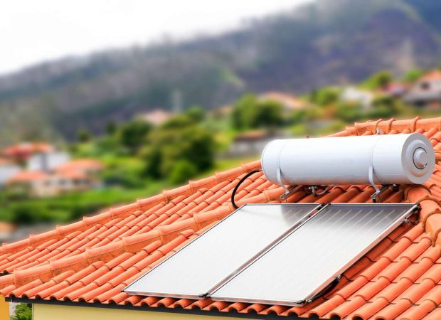 A close-up image of a solar water heater system installed on a red-tiled roof. The solar panels are positioned below a cylindrical water tank, with a scenic background of houses and greenery.